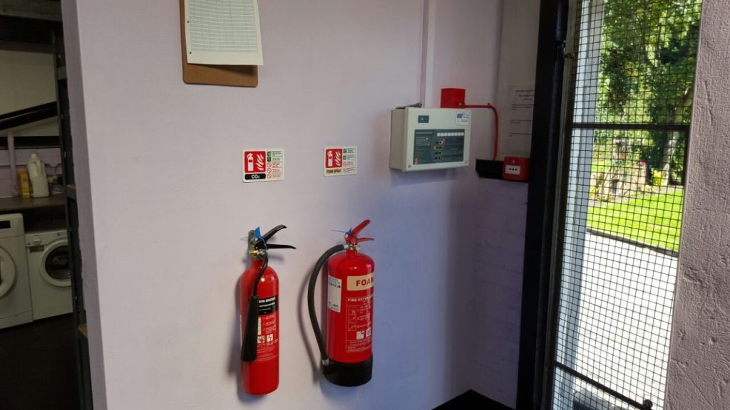 An indoor utility area featuring a fire safety station with two red extinguishers (CO2 and Foam) mounted on a purple wall next to a fire alarm control panel.