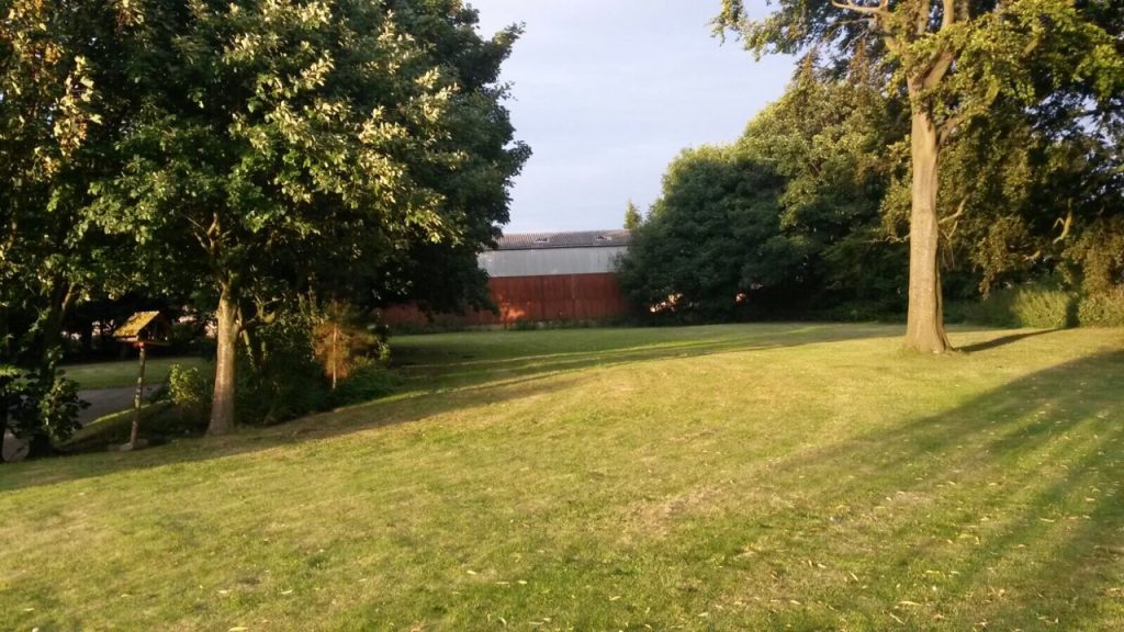 A sloping green lawn bordered by mature trees, with a large barn-like building with a red and white facade in the background.